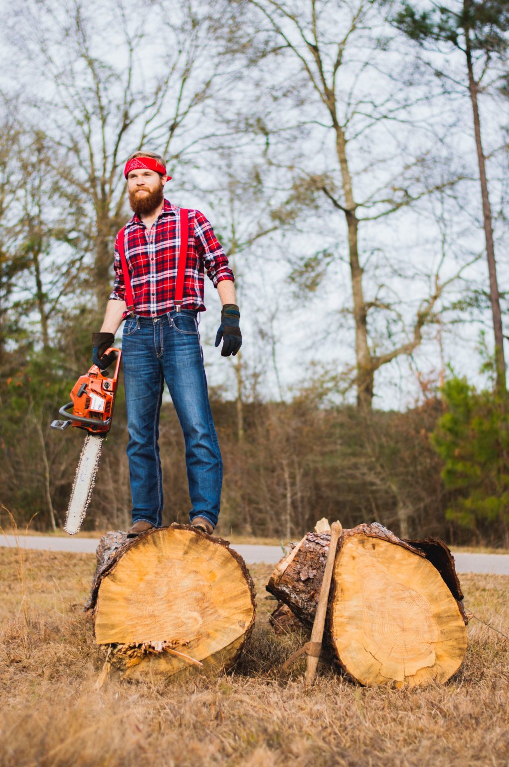 A bearded man in a red plaid shirt stands on a log, holding a chainsaw after cutting through wood.