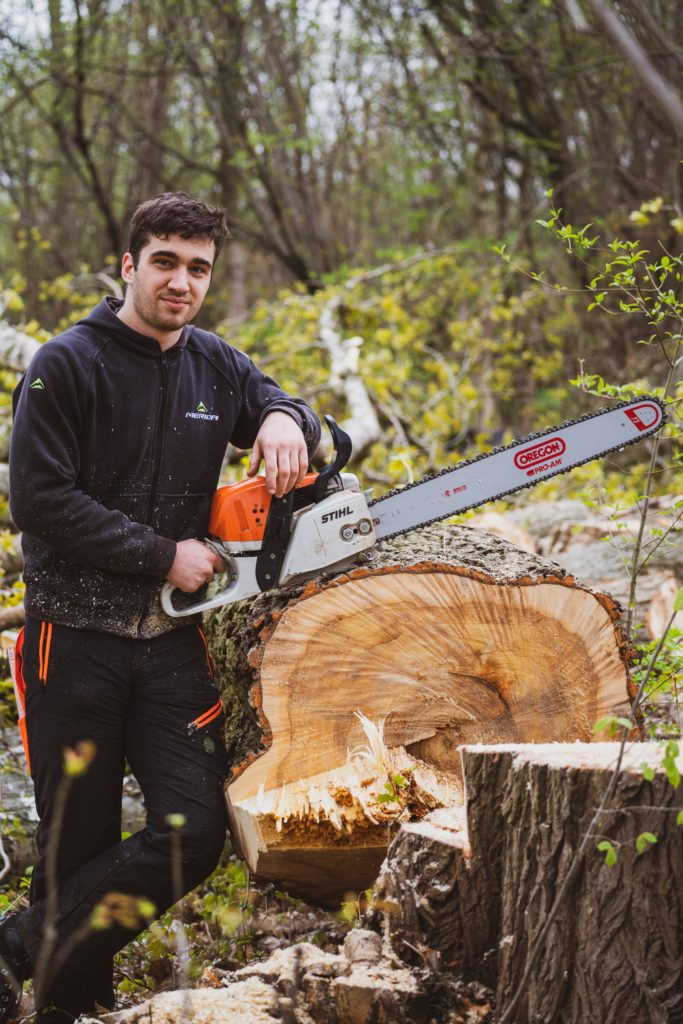 A young man dressed in black holds a chainsaw and leans against a huge log that he has cut.