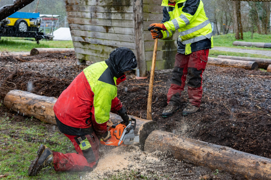 Young loggers working on a playground, cutting tree trunk with chain saws in rainy day.