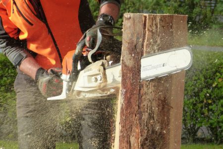 A man in an orange vest uses a standard chainsaw to make a vertical cut in a piece of wood.